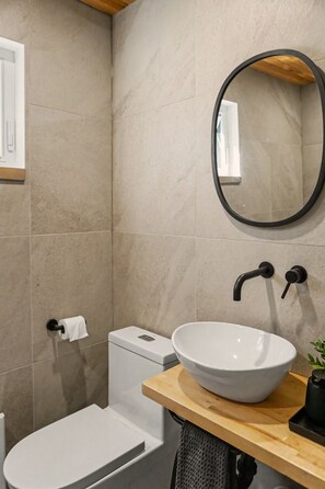 Un baño con inodoro blanco, lavamanos blanco con un recipiente grande y un espejo sobre el lavamanos.