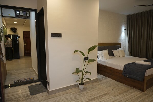 A hotel room with a bed, a potted plant, a wooden headboard, and a view of the hallway.