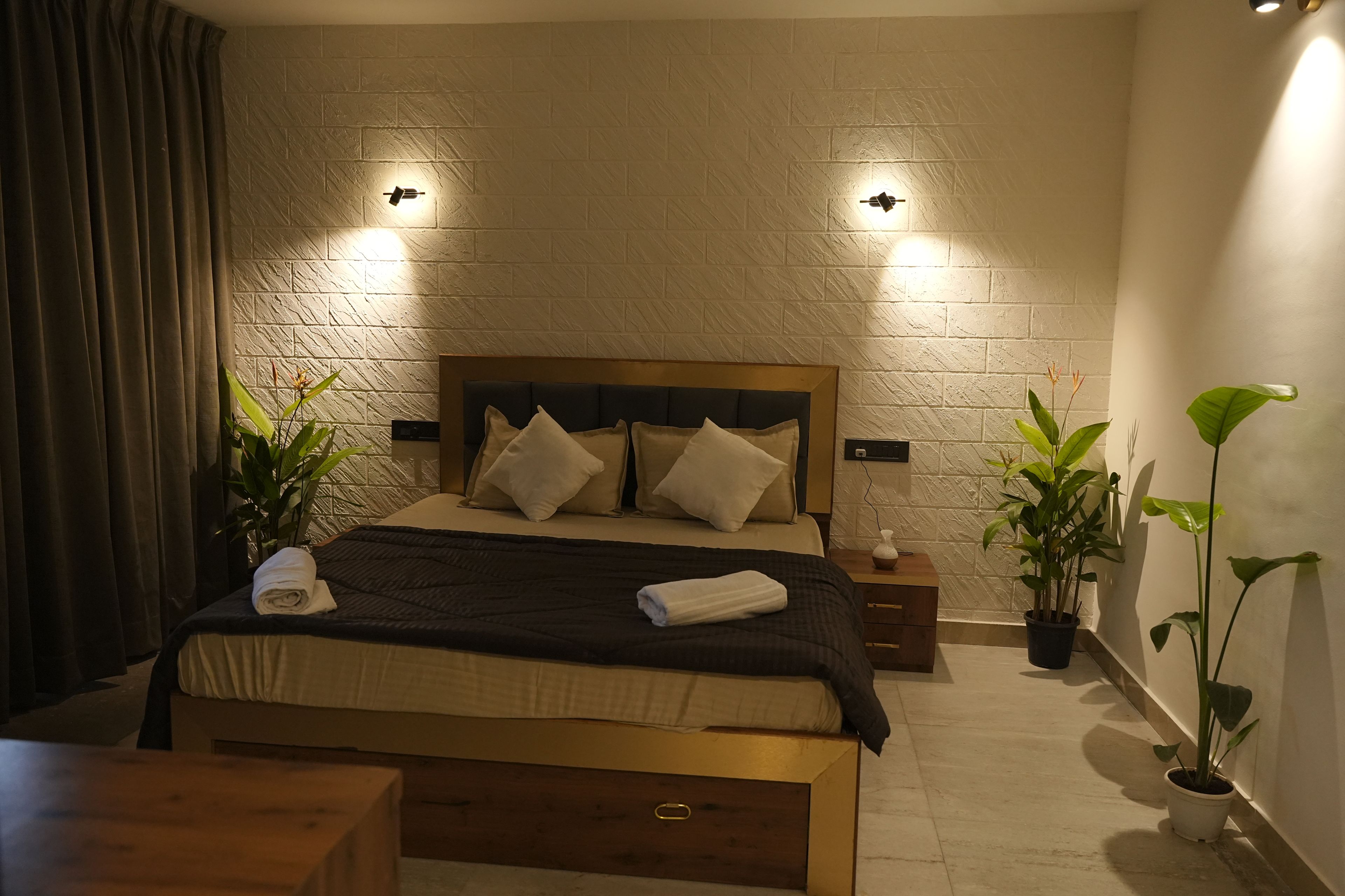 A hotel room with a brick-patterned wall, a bed with a wooden headboard, two bedside tables, a nightstand, a wooden bench, and two potted plants.
