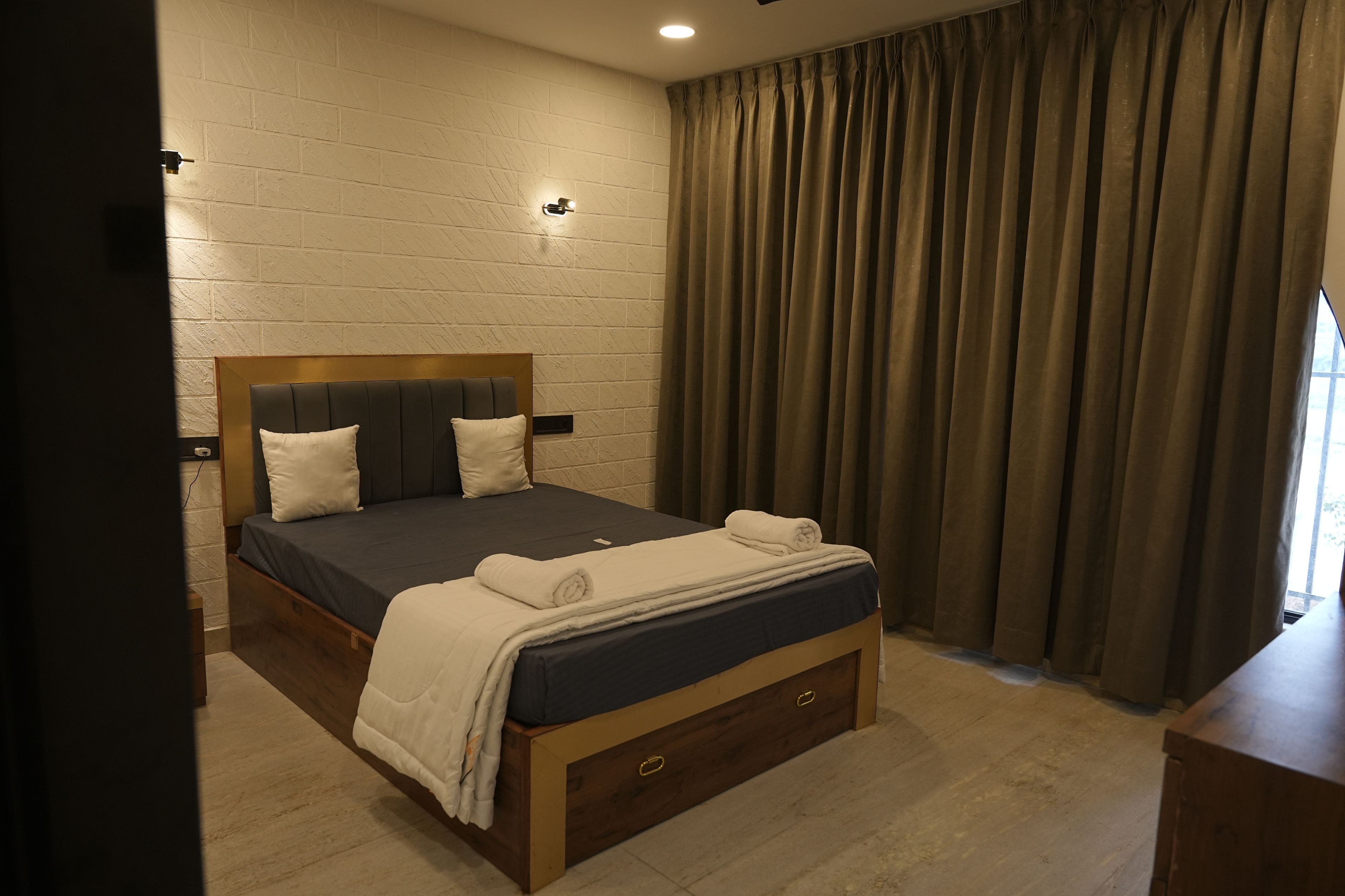 A bedroom with a wooden bed frame, a dark mattress, two pillows, a white blanket, and a wooden nightstand.