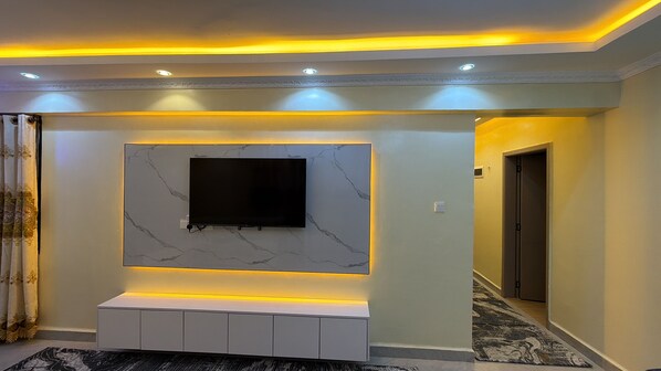 A modern living room with a flat-screen TV mounted on a marble wall, recessed lighting, and a white entertainment unit.