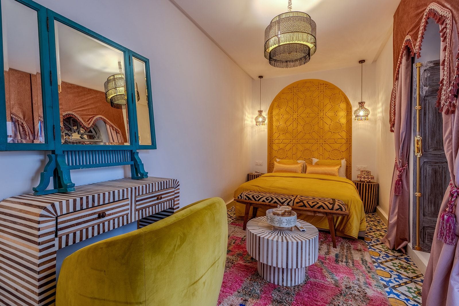 Ein Zimmer mit einem Bett, einem Spiegel, einer Holzkommode, einem runden Hocker und einer Pendelleuchte.