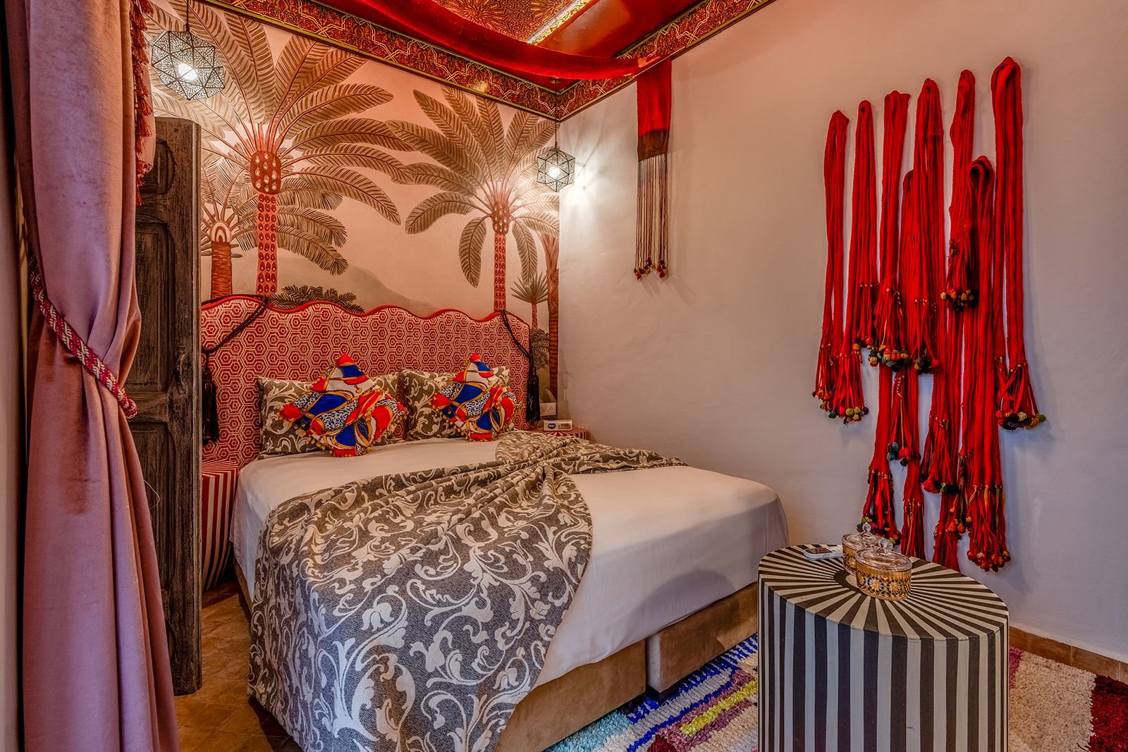 Een kamer met een bed, beddengoed met een patroon, een decoratief hoofdbord, een muur met rode franjes, en een klein nachtkastje met een gestreept voetenbankje.