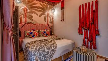 Een kamer met een bed, beddengoed met een patroon, een decoratief hoofdbord, een muur met rode franjes, en een klein nachtkastje met een gestreept voetenbankje.