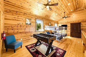 Ein gemütliches Zimmer mit Holzwänden, einem Deckenventilator, Etagenbetten, einem Tischfußballspiel, einem blauen Sofa und einer dunklen Holzkommode.