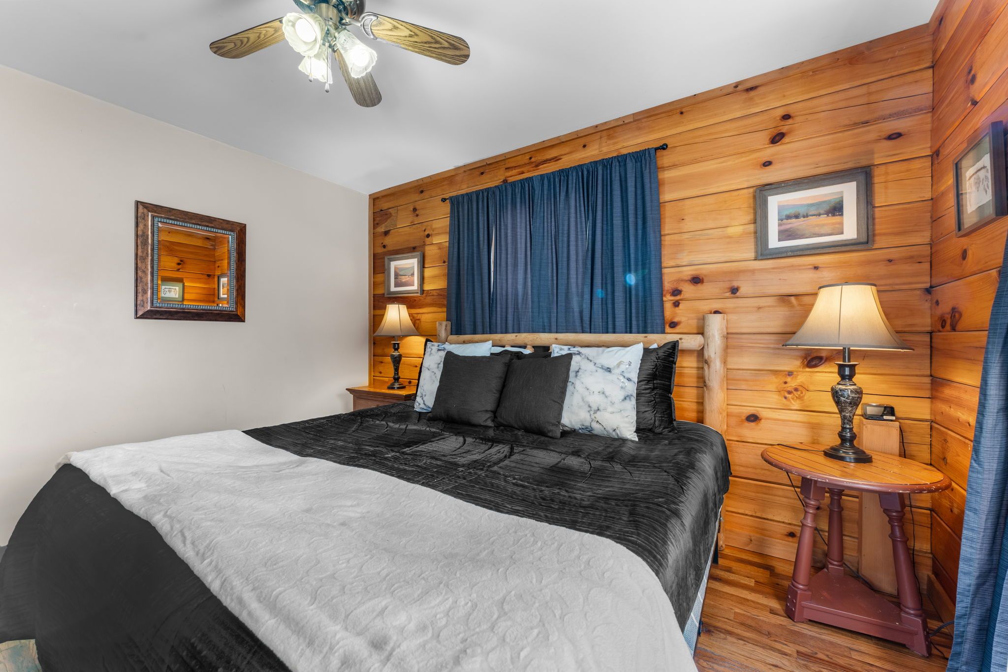 Un dormitorio con cama, dos mesitas de noche con lámparas, ventilador de techo, paredes de madera y un cuadro enmarcado en la pared.