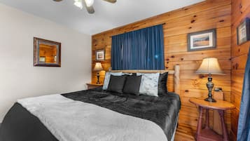 Un dormitorio con cama, dos mesitas de noche con lámparas, ventilador de techo, paredes de madera y un cuadro enmarcado en la pared.