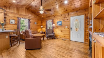 Interior de una cabaña con paredes de madera, ventilador de techo, zona de comedor con mesa y sillas, y una puerta con panel de vidrio decorativo.