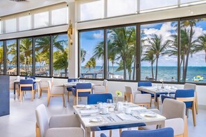 Ein Restaurant mit gedeckten Tischen, großen Fenstern und Blick auf Palmen und das Meer.