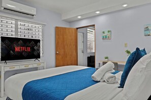 Una habitación de hotel con una cama, un televisor mostrando 'NETFLIX', una puerta de madera y un pequeño escritorio con una planta.