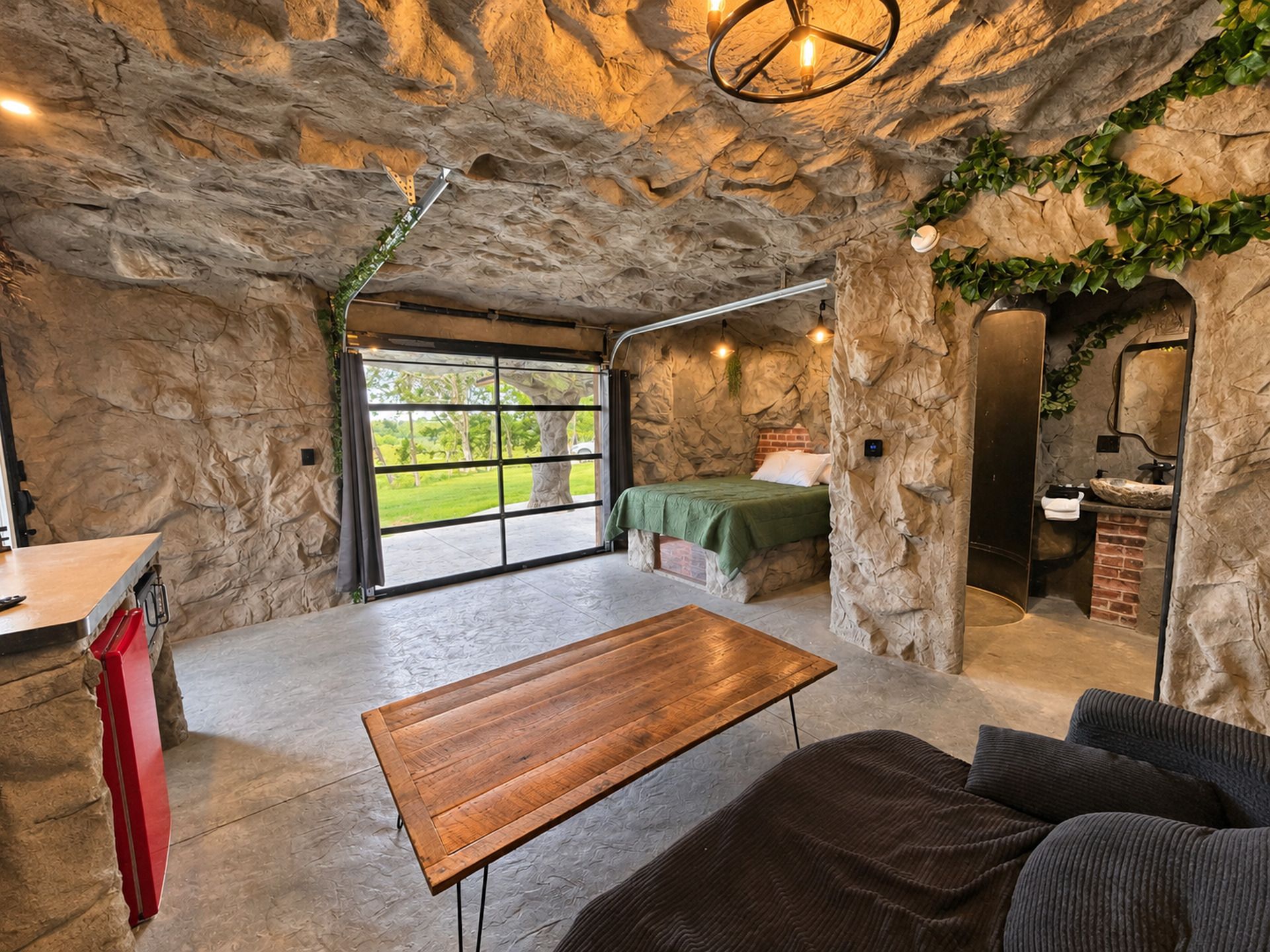 A room with a stone wall, a bed, a wooden table, a sofa, and a bathroom with a brick wall.