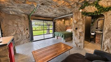 A room with a stone wall, a bed, a wooden table, a sofa, and a bathroom with a brick wall.