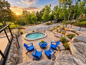 Un jacuzzi rodeado de sillas azules y un patio de piedra, con una terraza y barandilla de madera a la izquierda.