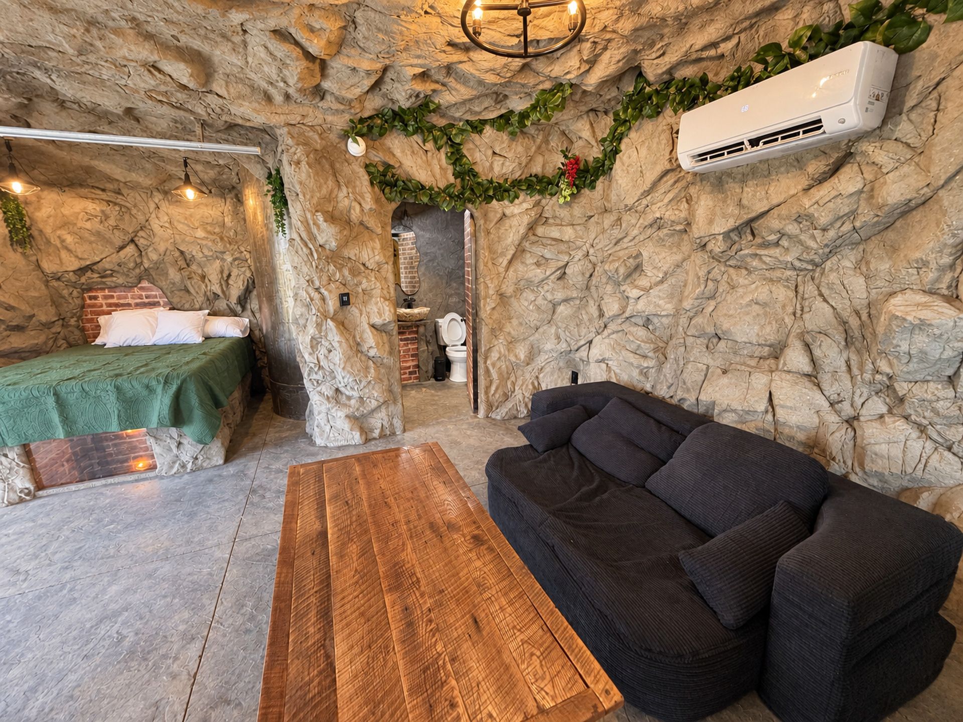 Una habitación con cama, sofá, mesa de madera y baño con pared de ladrillo.