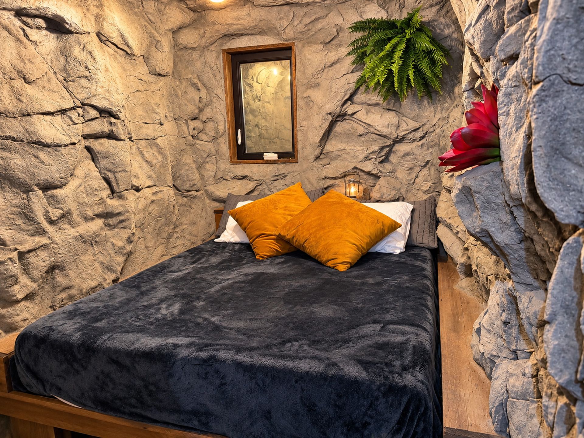 Una cama con una manta oscura y dos almohadas, un cuadro en la pared y una planta a la derecha.
