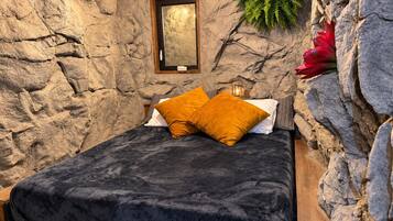 Una cama con una manta oscura y dos almohadas, un cuadro en la pared y una planta a la derecha.