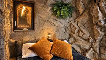 A bed with dark bedding and two brown pillows, a framed picture on the wall, and a small plant.