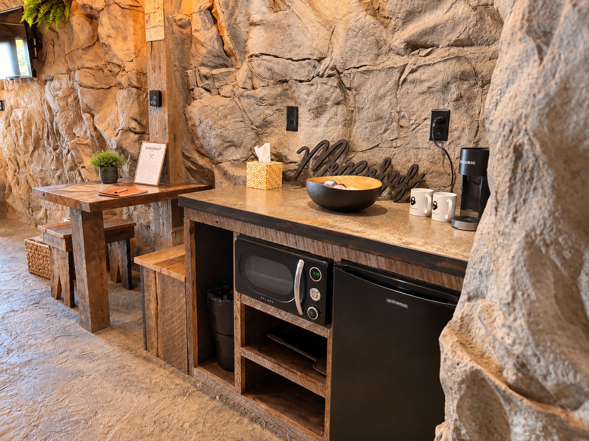 Una cocina con un fondo de pared de piedra, una mesa de madera, un tazón, una cafetera y un microondas.