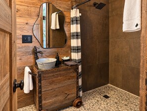 Un baño rústico con tocador de madera, un espejo redondo, una ducha con cabezal de ducha tipo lluvia negro y cortina de baño a rayas.