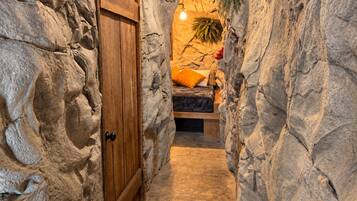 A narrow stone-walled corridor leading to a bedroom with a bed, a nightstand, and a hanging plant.