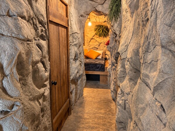 Un estrecho pasillo de paredes de piedra que conduce a un dormitorio con una cama, una mesita de noche y una planta colgante.