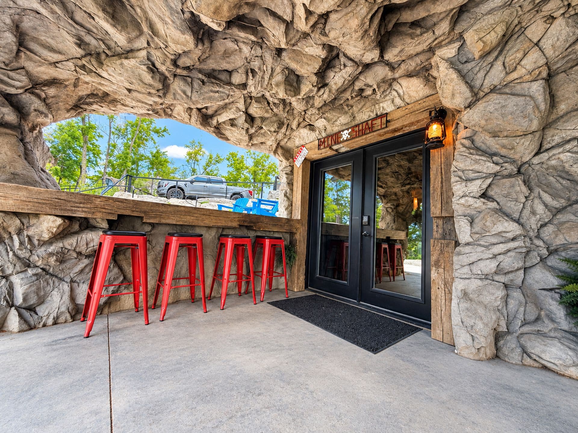 A restaurant entrance with a stone facade, outdoor seating with red stools, and a car parked outside.