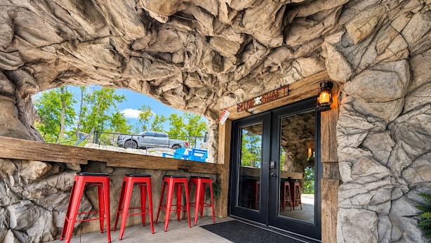 Entrada de restaurante com fachada de pedra, mesas ao ar livre com bancos vermelhos e um carro estacionado na frente.