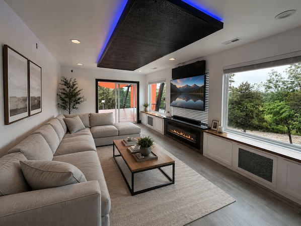 A modern living room with a sectional sofa, a fireplace, and a large window with a view of greenery. - Altura Tower - With a View (Lancaster)