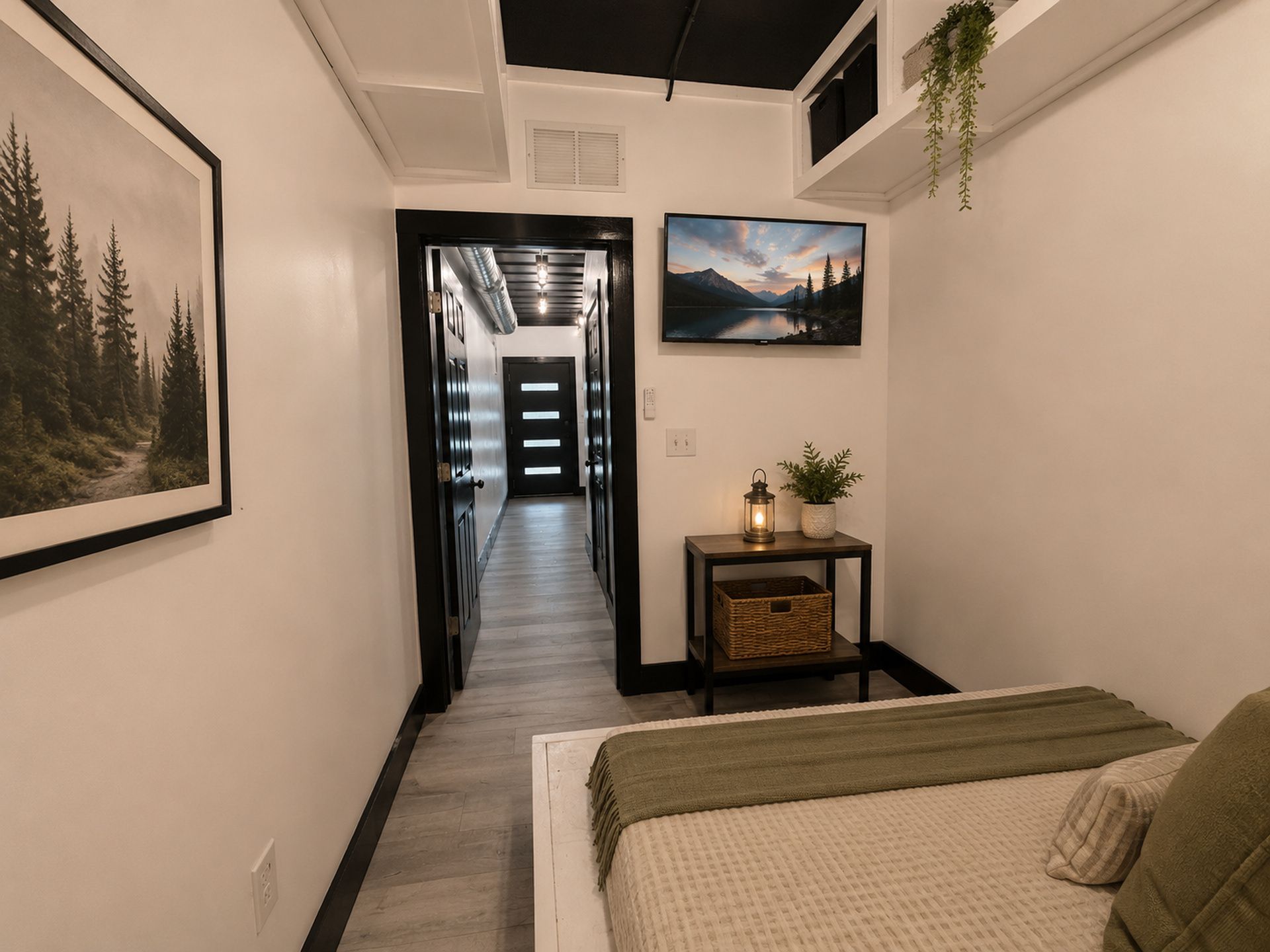 A hotel corridor with a bed, a framed picture, a small table with a lamp, and a hanging plant.