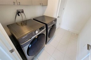 Een wasserette met twee Samsung wasmachines en een droger, een vloer met witte tegels en een deur aan de rechterkant.