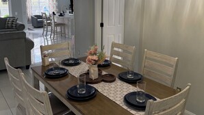 Un coin repas avec une table en bois dressée pour six personnes, entourée de chaises blanches, sous un lustre en cristal.