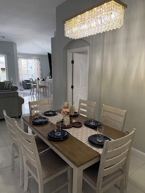 A dining area with a wooden table set for six, surrounded by white chairs, under a crystal chandelier. - Unique Home Gated Community Heated Pool 5 miles to Disney (Kissimmee)