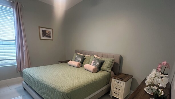 Une chambre à coucher comprenant un lit, des tables de chevet, un ventilateur de plafond, une fenêtre avec des rideaux et une petite table de nuit sur laquelle est posée une plante en pot.