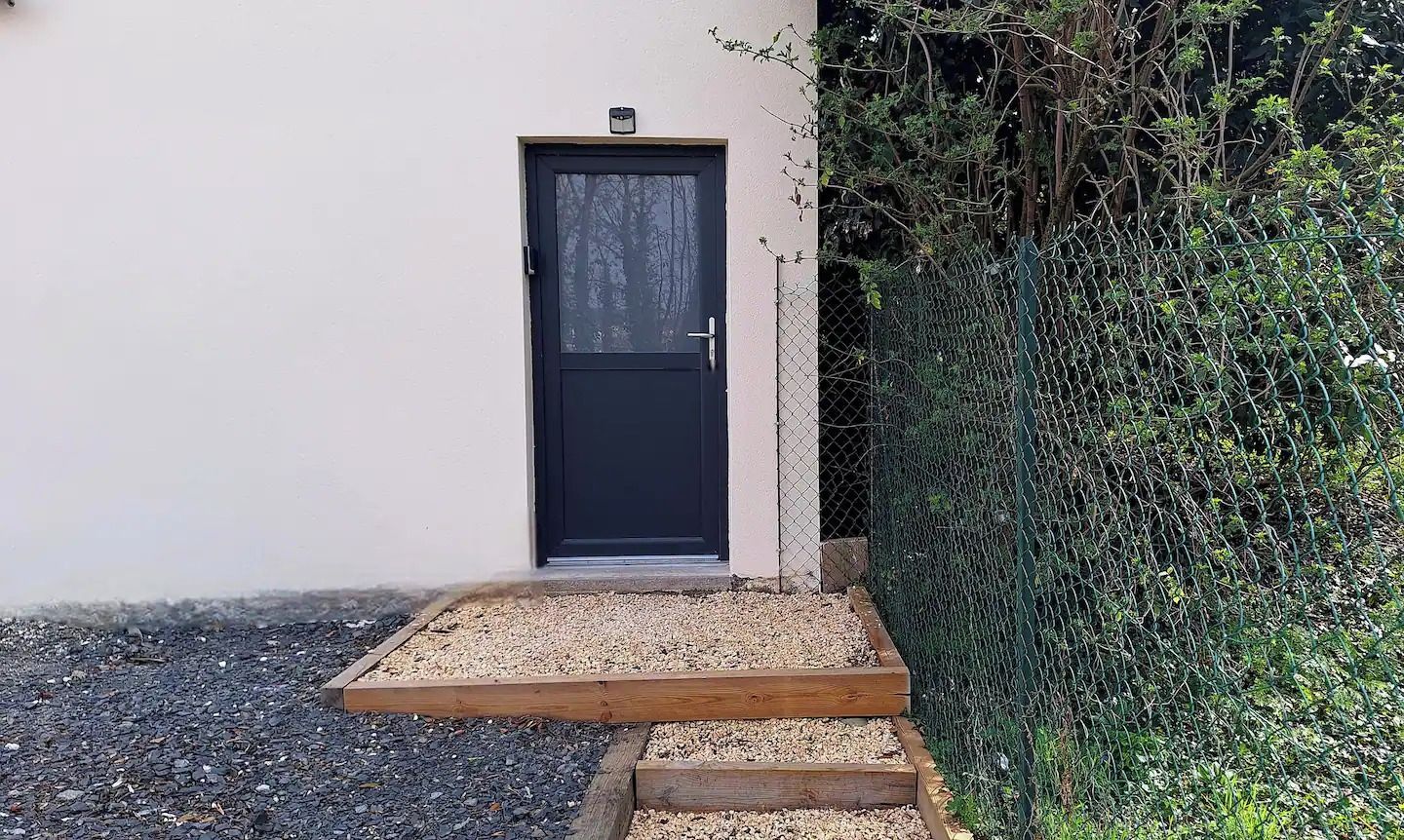 Une porte noire, précédée d’un petit seuil, entourée de gravier et bordée de bois.