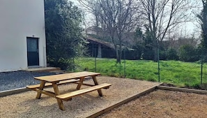 A picnic table in a garden area with a gravel path, surrounded by a fence and trees. - Two-bedroom apartment with outdoor space and a pétanque court (Pérignat-sur-Allier)