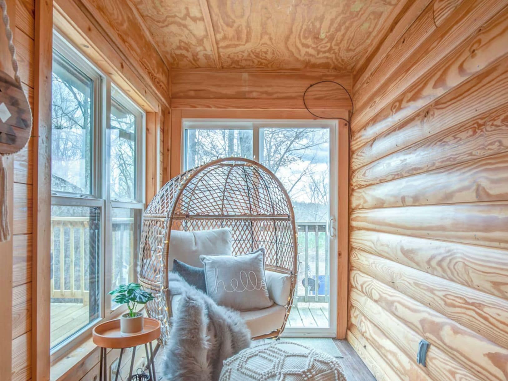 Un intérieur de chalet chaleureux avec une façade en bois, une grande fenêtre, un fauteuil suspendu, une petite table avec une plante et un tapis moelleux.