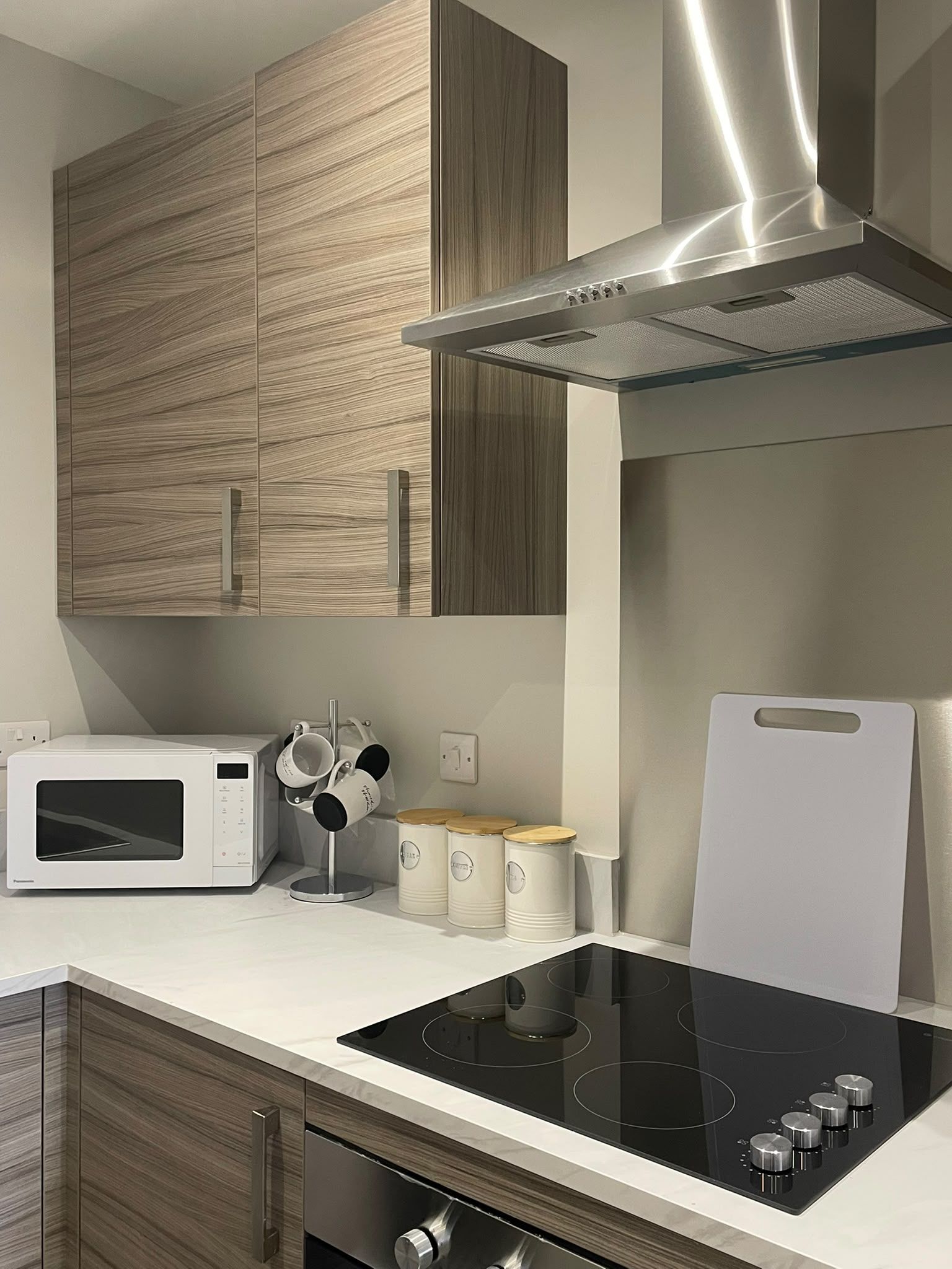 A modern kitchen with a built-in microwave, a stovetop with a glass cooktop, wooden cabinets, and a stainless steel range hood.