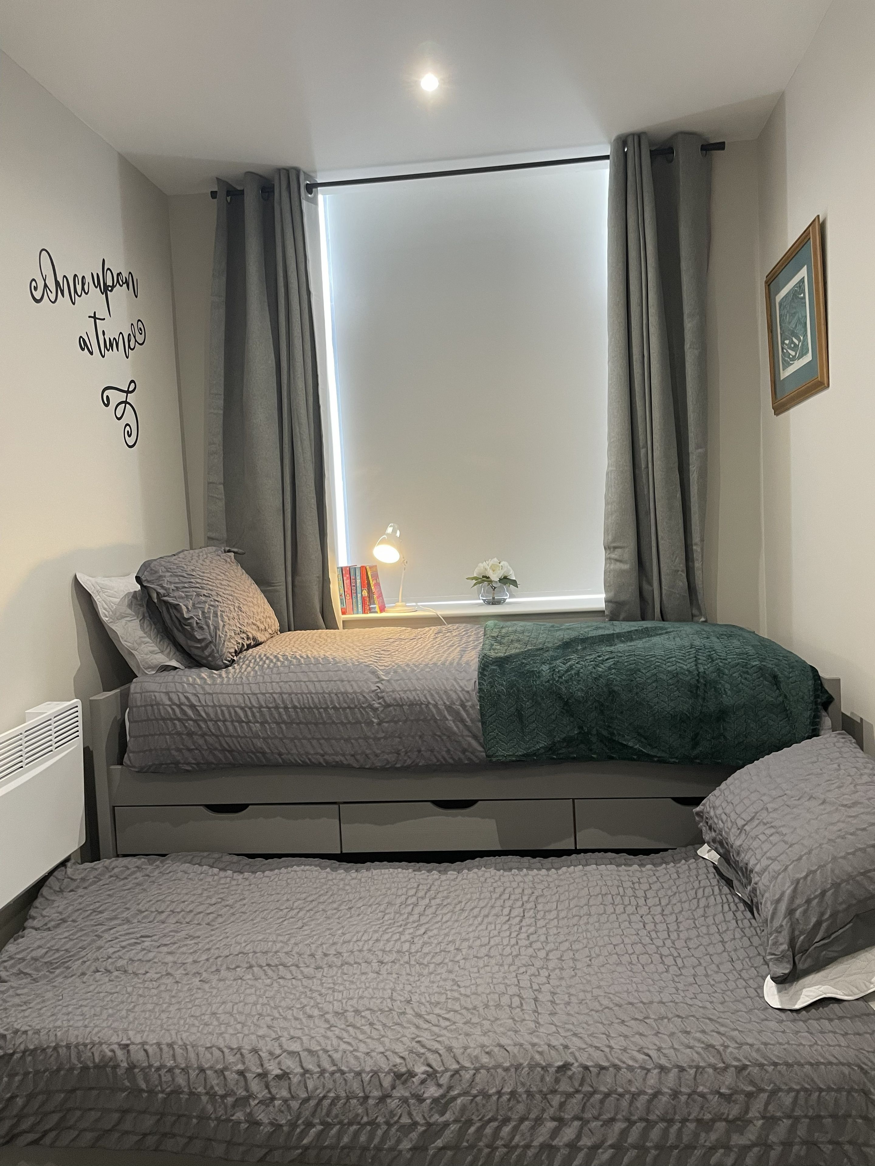 A bedroom with a bed, a bedside table with a lamp, a window with curtains, and a wall decoration.