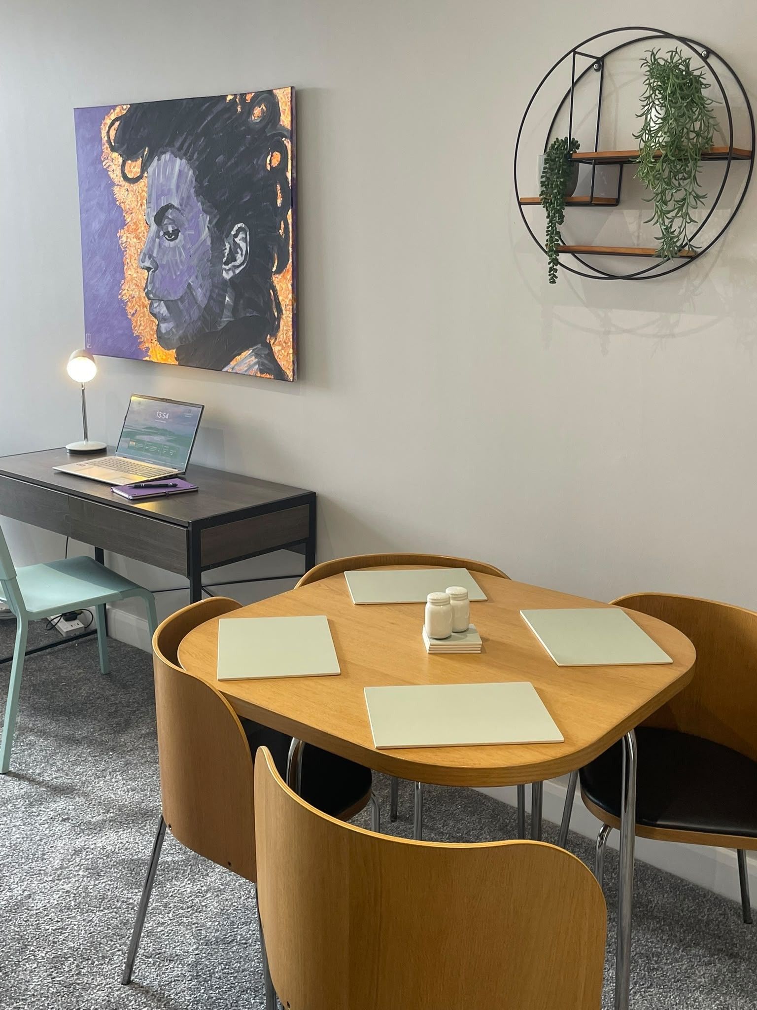A modern office space with a round table, two chairs, a desk with a laptop, a painting, and a wall-mounted plant.