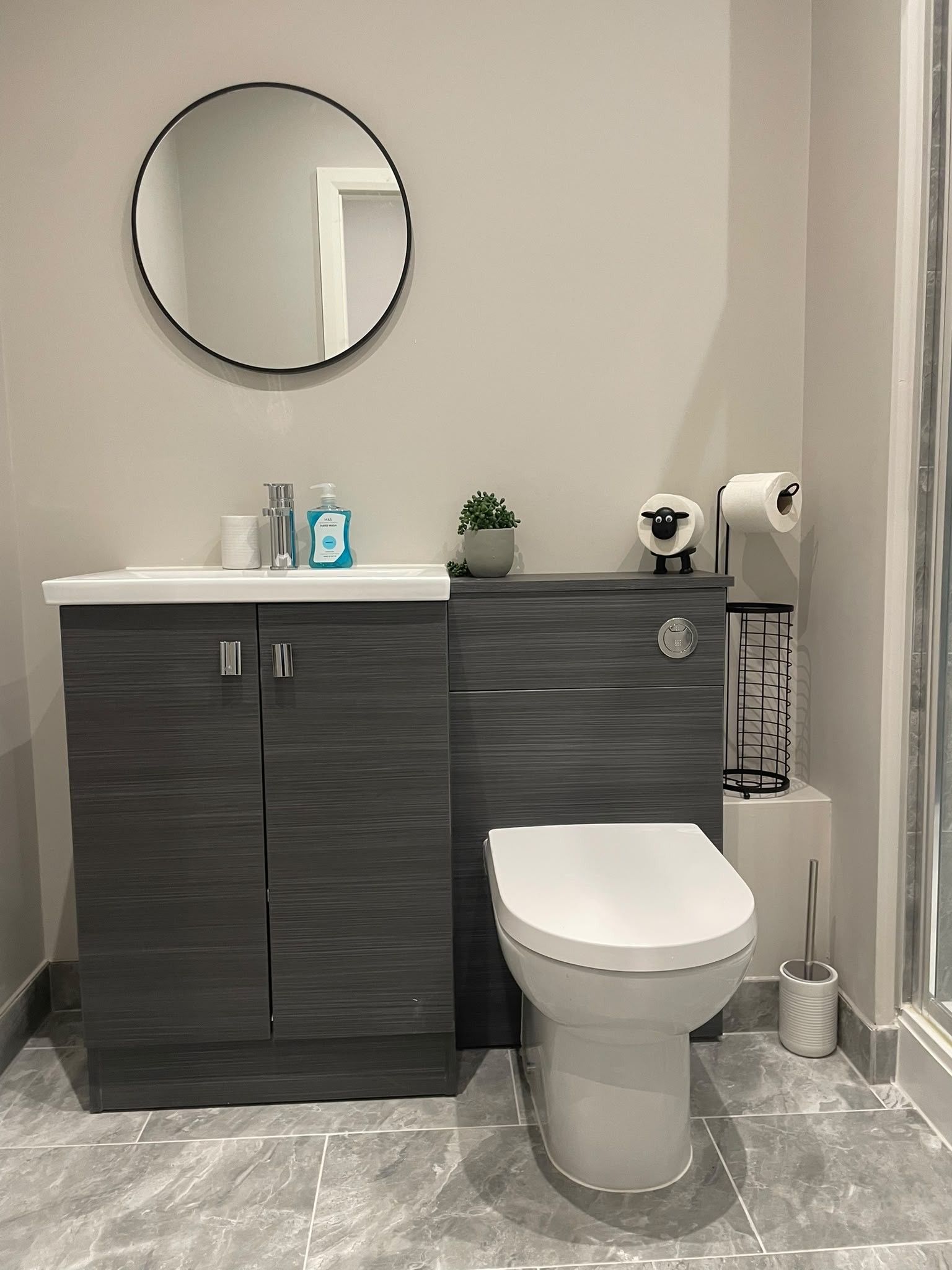 Ein Badezimmer mit dunklem Holzschrank, einer weißen Toilette, einem runden Spiegel und einem kleinen Topfpflanzen.