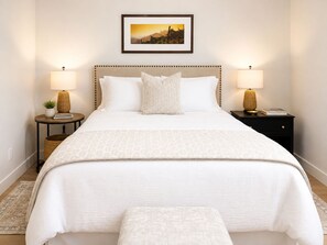 A neatly arranged bedroom with a bed, bedside tables, lamps, a framed picture on the wall, and a small potted plant. - Westshore Oasis | 1-Bedroom Victoria Retreat (Victoria)