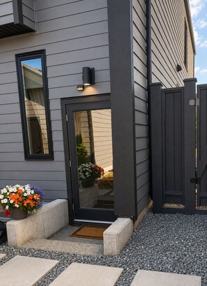 A modern house with a grey siding exterior, a black door, and a small garden with potted plants. - Westshore Oasis | 1-Bedroom Victoria Retreat (Victoria)
