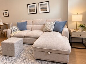 A beige sectional sofa with a matching ottoman, a small side table, a lamp, and framed artwork on the wall. - Westshore Oasis | 1-Bedroom Victoria Retreat (Victoria)