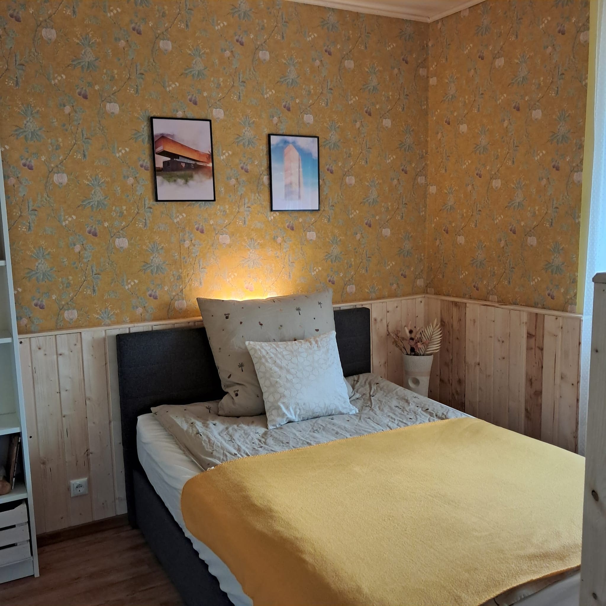 A bedroom with a bed, a nightstand, a wall-mounted lamp, and two framed pictures.