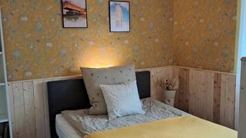 A bedroom with a bed, a nightstand, a wall-mounted lamp, and two framed pictures.