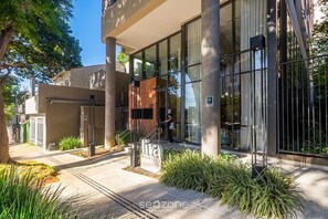 A modern building entrance with glass doors, a signboard, and a person standing by the entrance. - Studio in Condo. w/ City View & Gym Ipr0410 (São Paulo)