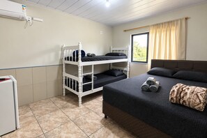 Una habitación con literas, una cama, un ventanal con cortinas y un electrodoméstico pequeño y blanco.