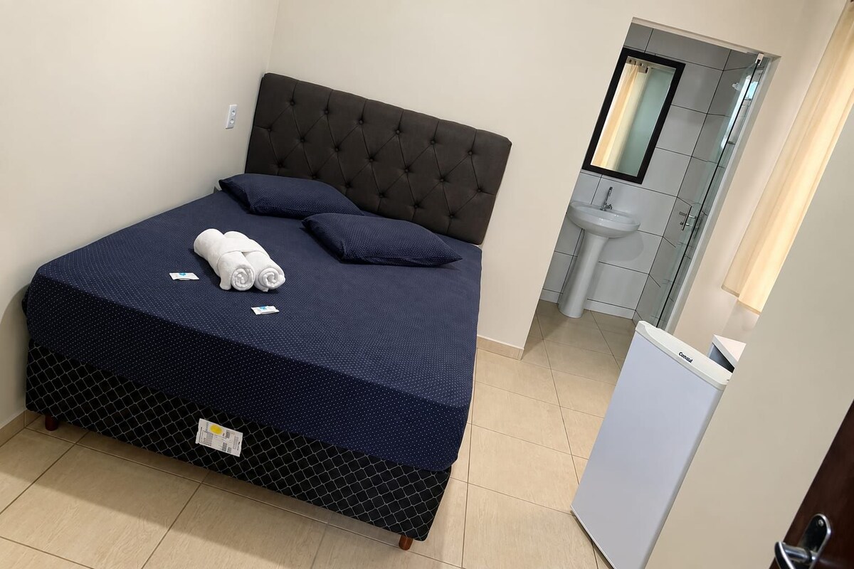 A neatly made bed with a dark headboard, a small bedside table, and a white sink in the adjacent bathroom.