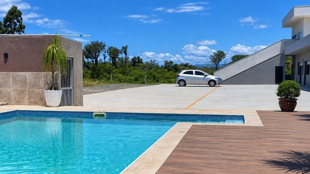 A swimming pool with a tiled edge, a paved deck, and a car parked in the background. - Pousada Tio Tonho (Penha)