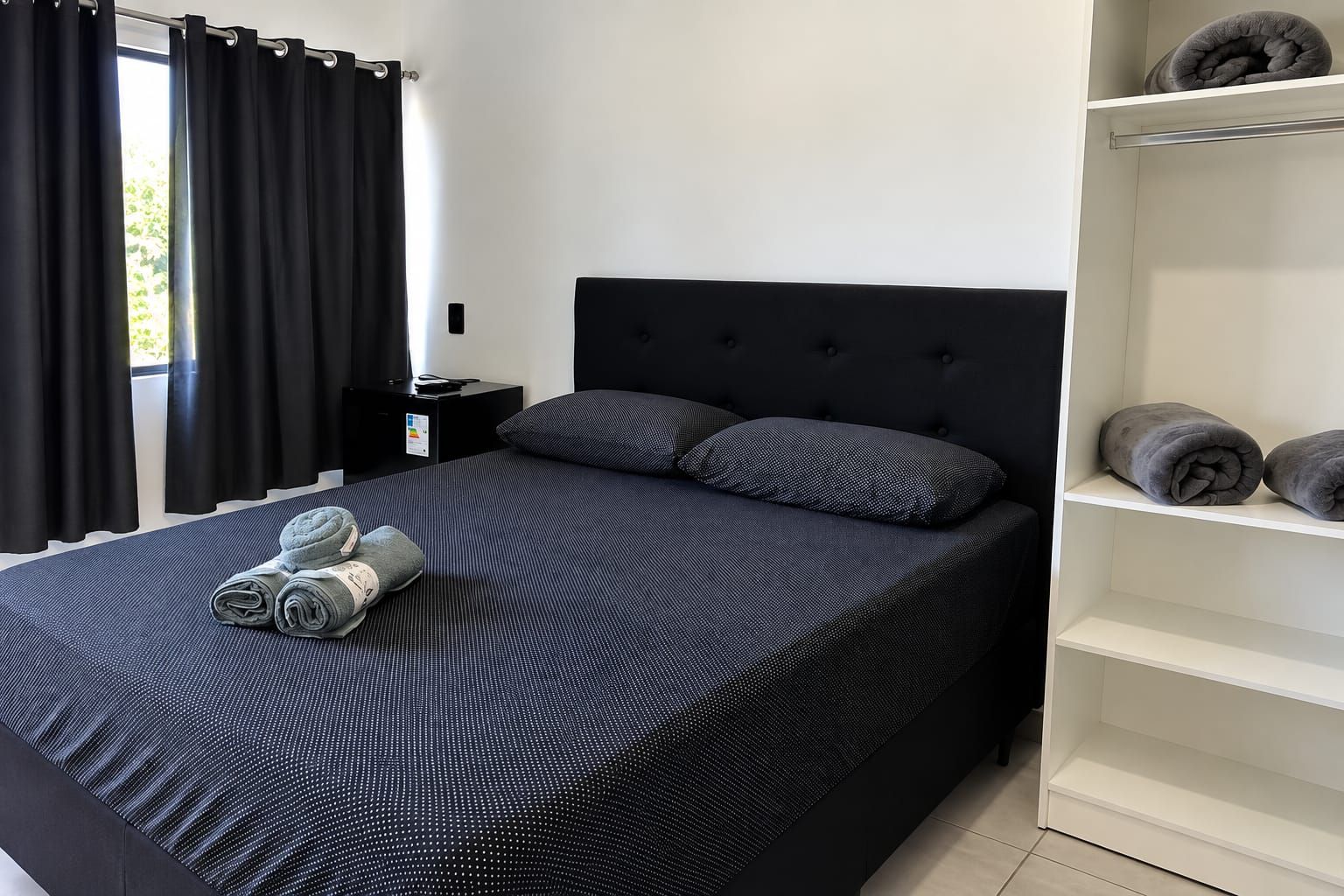 Ein Schlafzimmer mit einem Bett, Kissen, einem Nachttisch, einem Regal mit Handtüchern und einem Fenster mit Vorhängen.